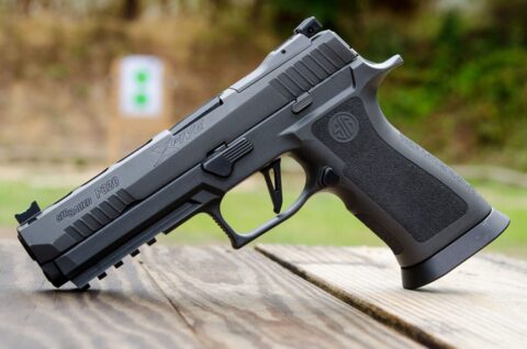 A Sig Sauer Legion P320 XFive pistol displayed on a dark surface. The firearm features a tungsten-infused grip module, a distinct gray PVD finish, and an extended barrel. The slide is precision-machined with lightening cuts, and the pistol is equipped with a flat-faced trigger, enhancing its accuracy and control. The Legion medallion is visible on the grip, indicating its elite status within the Sig Sauer Legion Series